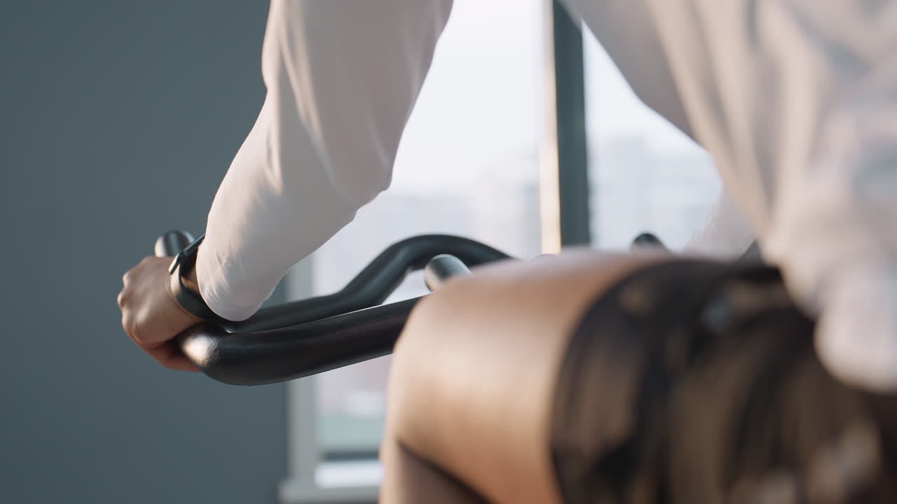 close up of boy hand gripping stationary bike handlebars in bright gym light filtering through large window, blurred urban buildings visible outside