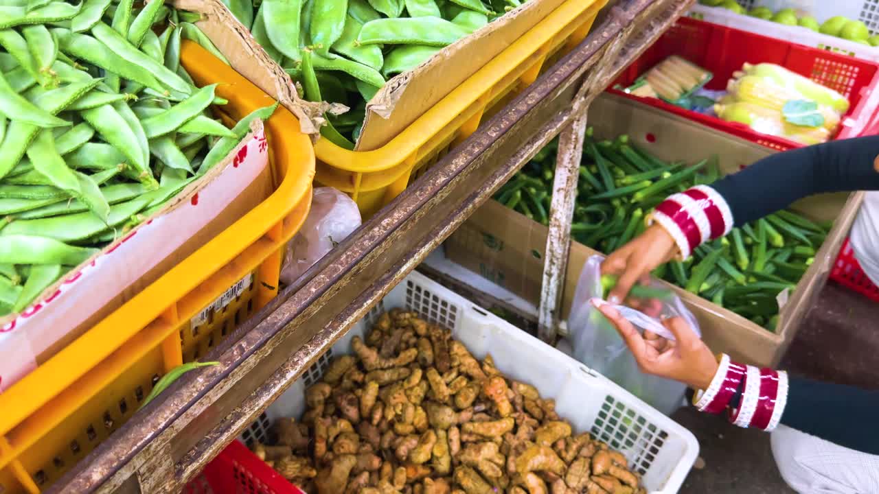 A person with striped sleeves chooses and bags fresh green beans and turmeric roots at a vibrant outdoor vegetable market, under natural daylight with a handheld camera angle
