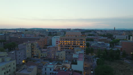 Forwards-fly-above-city-at-twilight.-Illuminated-contemporary-multistorey-apartment-building-or-hotel-with-rooftop-terrace.-Rome,-Italy