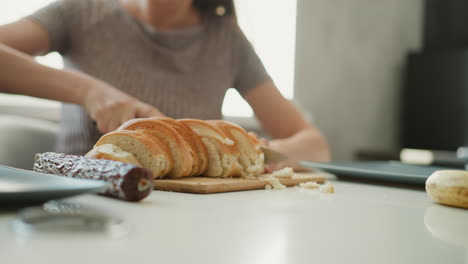 close up of baker slicing loaf of bread with knife on wooden board focus on serrated blade cutting through crust into even slices with crumbs falling on white table under soft window light in kitchen