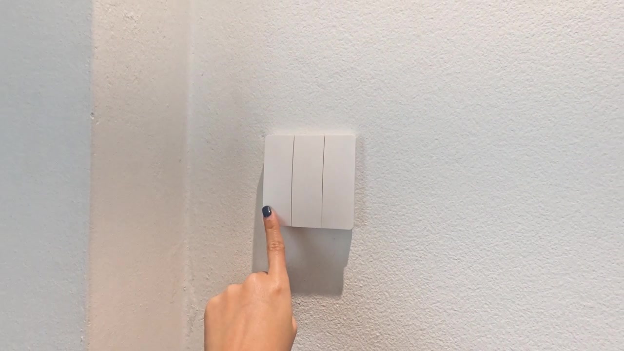 A finger toggles a white light switch on a wall, adjusting indoor room lighting