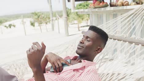 happy african american man using smartphone lying in hammock on sunny beach, slow motion