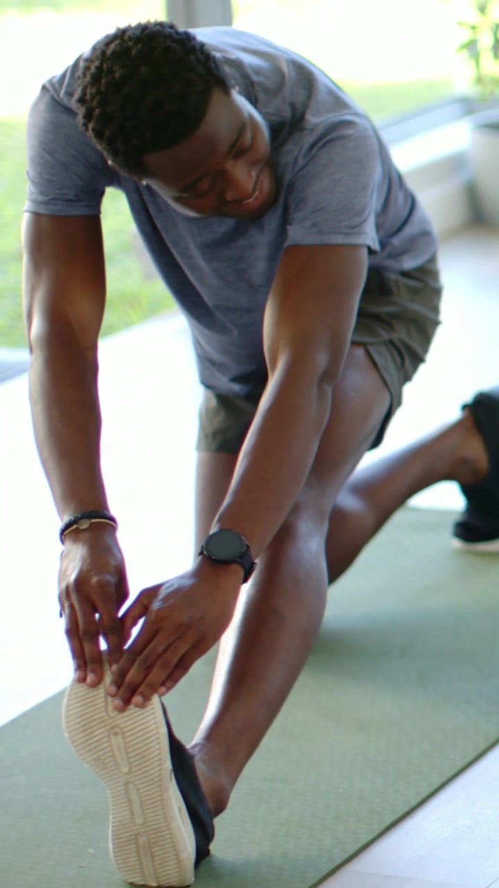 Vertical video: Entering left, man in gray tee stretching in lunge on mat with bottle, warming up