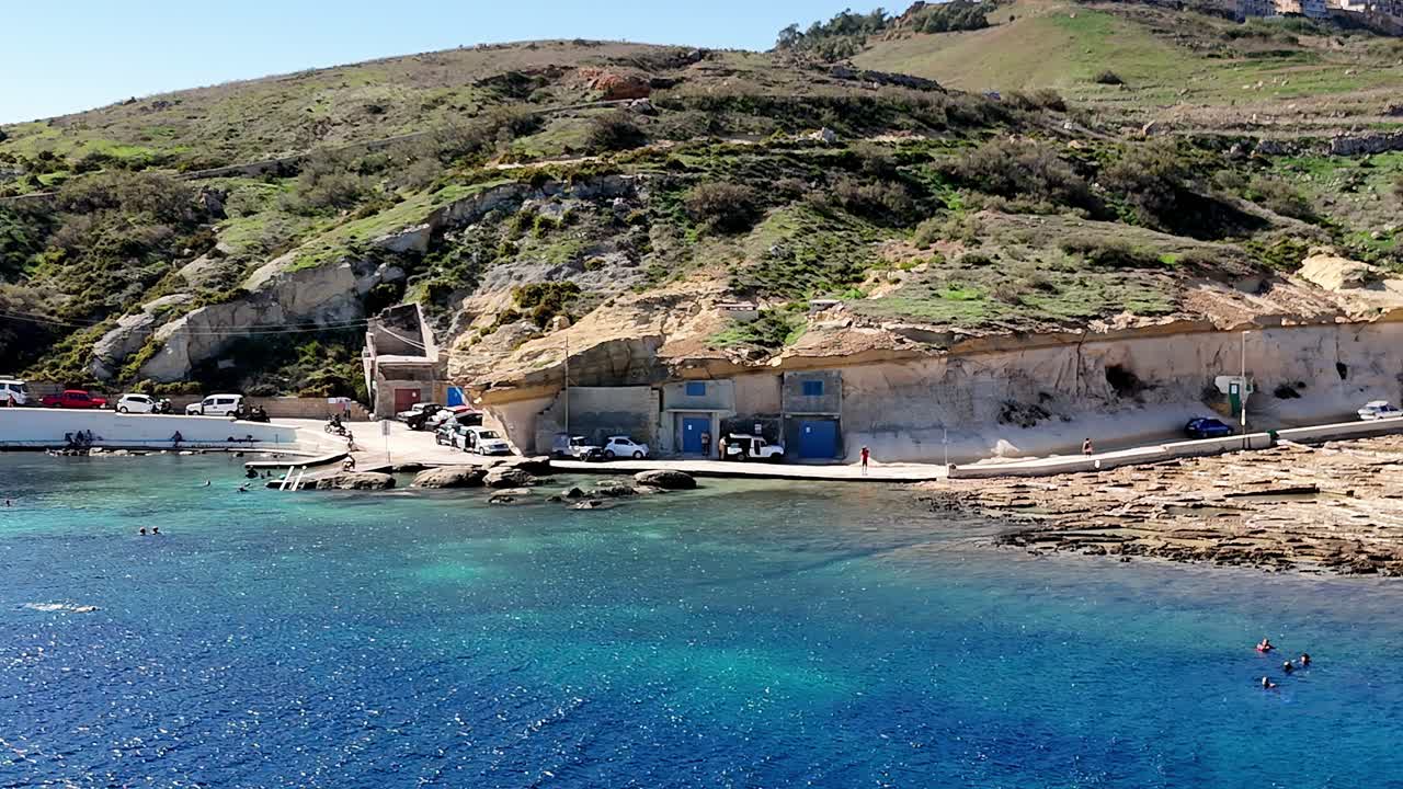 Yüksek bir açıdan drone ile çekilen bu fotoğrafta, Malta, Gozo'daki Daħlet Qorrot Koyu'nun engebeli kireçtaşı kayalıkları, parlak yaz güneşinin altında kayaların içine yerleşmiş canlı mavi kapılı kayıkhane evlerini gözler önüne seriyor.
