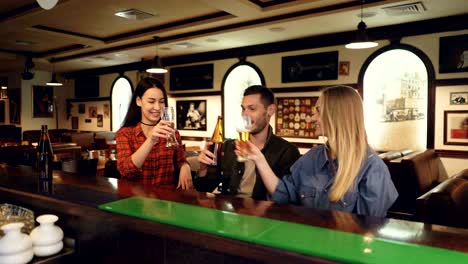 two attractive young women and bearded young man are drinking beer sitting at bar counter, clinking and talking. friends meeting in pub