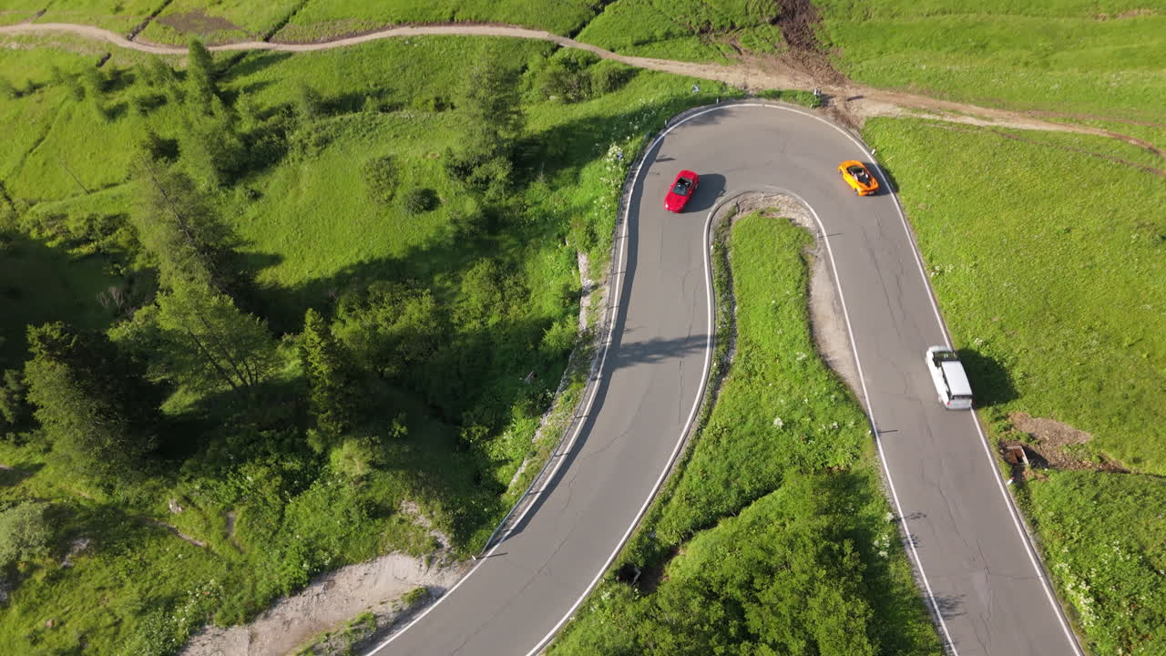 Drone aerial shot of luxury sports cars entering a sharp hairpin turn on a mountain serpentine road. Scenic alpine landscape. Category: Transportation