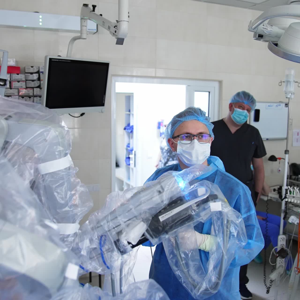 Male doctor in glasses and cap cooperates with modern robotic surgeon. Medical team watches the process of operation at the screen