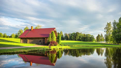 Timelapse-shot-of-a-wooden-cottage-with-white-clouds-passing-over-green-grasslands-alongside-a-small-lake-on-a-cloudy-day