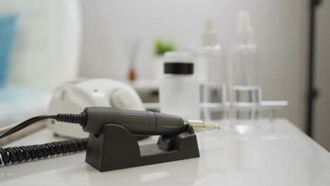 view of professional salon table with spray bottles, lotion dispenser, and electric nail drill arranged neatly on white surface with blurred background featuring indoor plant
