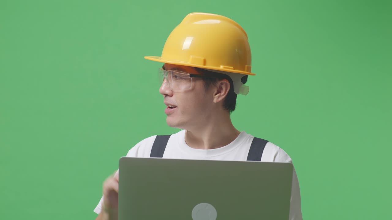녹색 화면 배경 스튜디오에 서 있는 동안 노트북을 사용하고 을 가리키는 안경과 안전 헬을 착용한 아시아인 노동자의 근접 사진