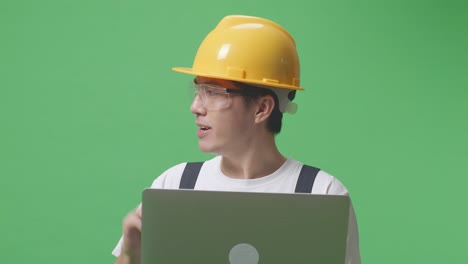 close up de un hombre asiático que trabaja con gafas de protección y casco de seguridad usando una computadora portátil y apuntando hacia un lado mientras está de pie en el estudio de fondo de la pantalla verde