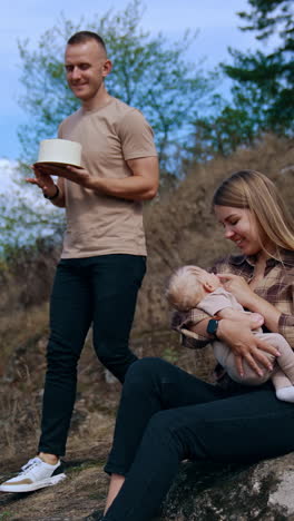 long-haired caucasian woman sits on the rock with her baby son in hands. man with a cake goes down the hill to join them. vertical video