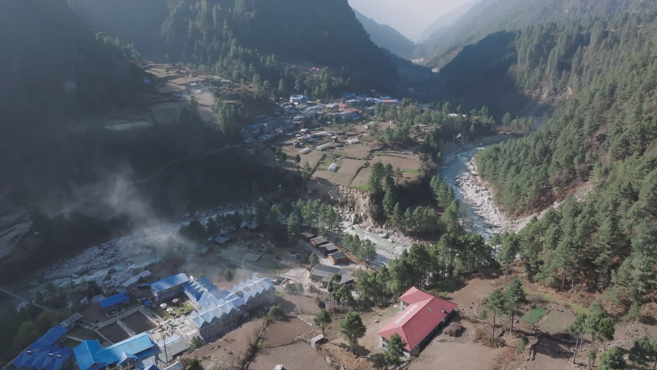 Drone shot of Phakding Village in early morning, sunlit houses, riverbank, misty clouds, forest backdrop, serene travel and tourism vibe in Nepal serenity