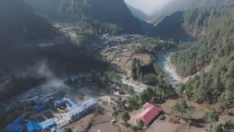 drone shot of phakding village in early morning, sunlit houses, riverbank, misty clouds, forest backdrop, serene travel and tourism vibe in nepal serenity