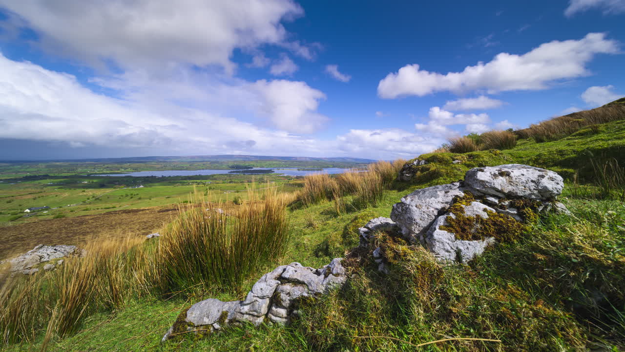 Zeitrafferaufnahme einer ländlichen Tal-Landschaft mit Felsformation im Vordergrund und vorüberziehendem Regensturm, Hügeln und See in der Ferne an einem Frühlingstag, aufgenommen von Carrowkeel in County Sligo in Irland