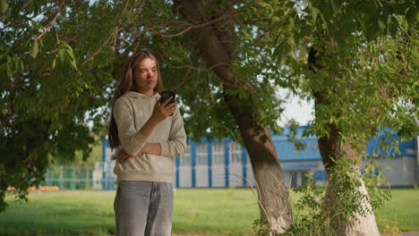 young woman intently focused on her phone under tree with sunlight creating soft shadows, she appears engaged with screen, surrounded by nature with building in background