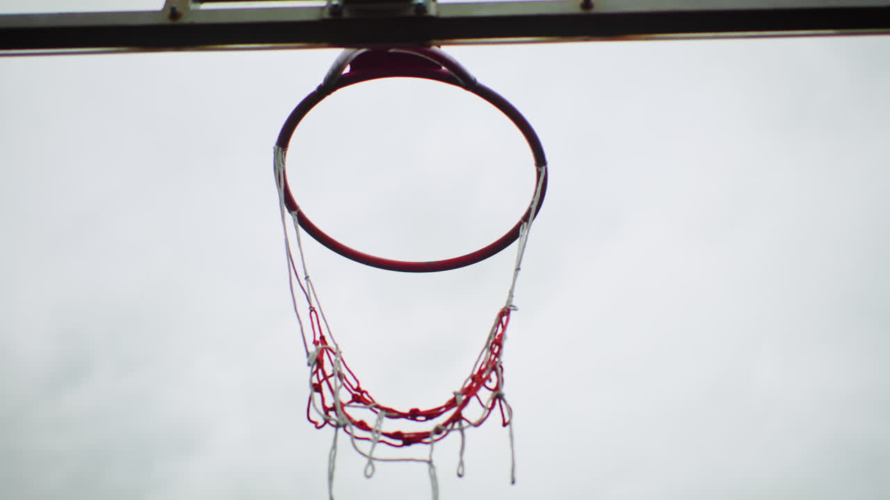 Canestro da basket vuoto incorniciato da nuvole, catena arrugginita e rete sfilacciata appese contro un cielo nuvoloso, campo silenzioso e deserto, atmosfera malinconica, tabellone texturizzato, composizione minimalista contemplativa