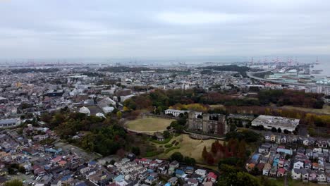 A-sprawling-cityscape-with-green-spaces-and-industrial-backdrop,-cloudy-sky,-aerial-view