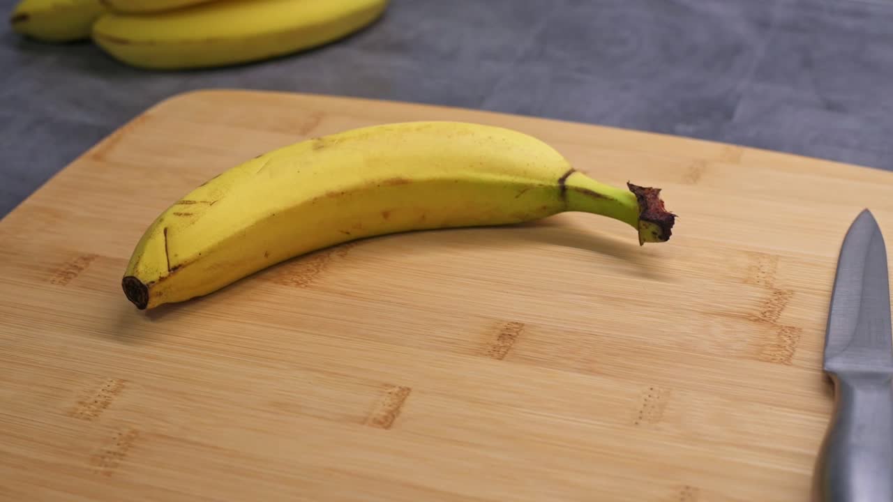 A ripe banana with slight blemishes rests on a wooden cutting board. A knife is placed nearby, suggesting preparation for slicing. The background features additional bananas on a dark countertop.