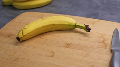 a ripe banana with slight blemishes rests on a wooden cutting board. a knife is placed nearby, suggesting preparation for slicing. the background features additional bananas on a dark countertop.