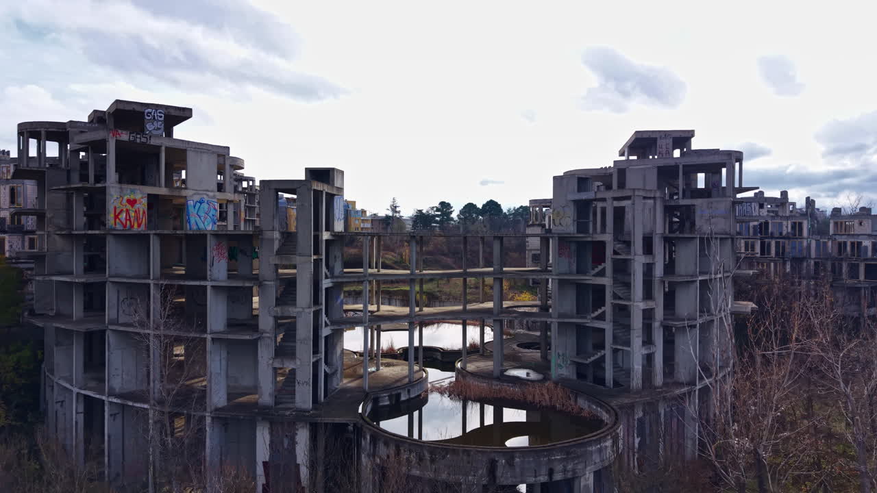 Zrujnowana architektura stoi upiornie cicho wokół spokojnego stawu na opuszczonej inwestycji, przywołując wspomnienia dawno zapomnianych marzeń pod ponurym niebem.
