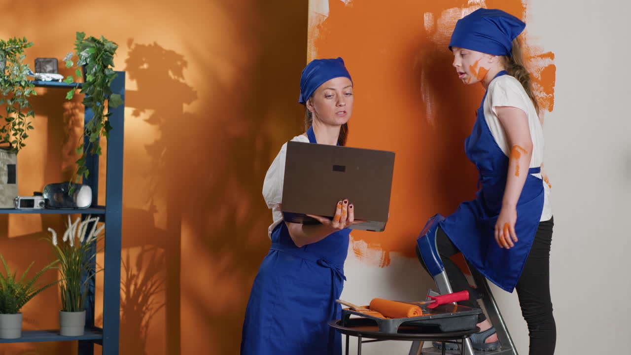 Mother and daughter painting a wall orange while using a laptop