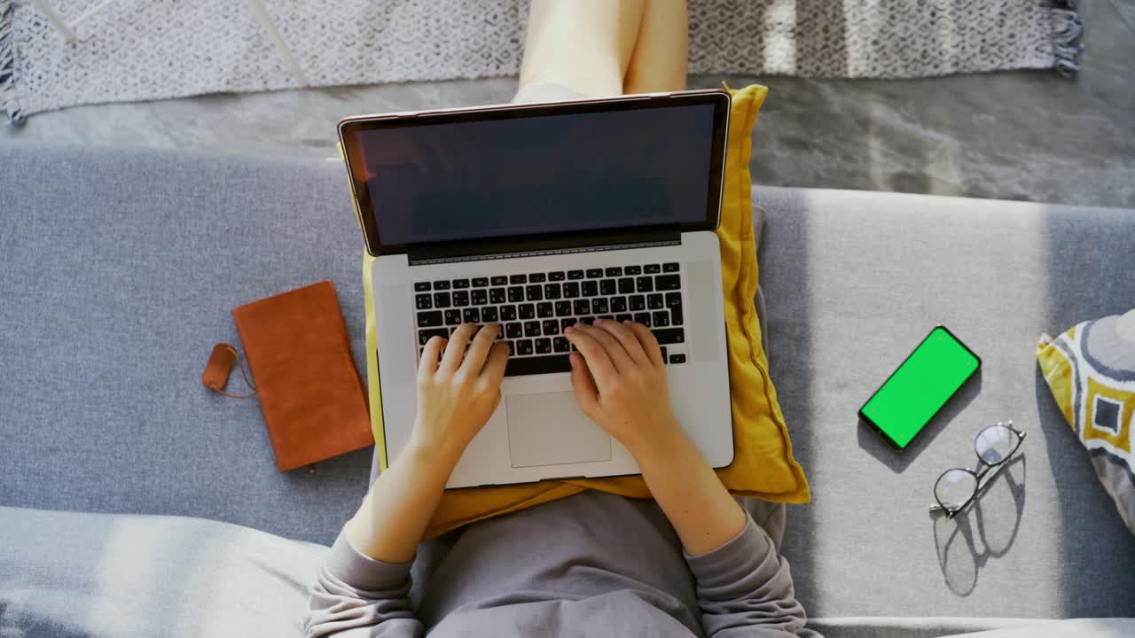 Woman types on laptop sitting on couch near mobile phone