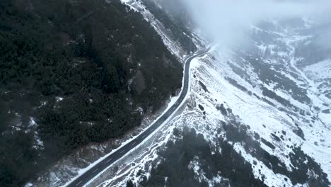 aerial view of a small car driving on a snow covered mountain road during a cloudy winter day with snowfall in switzerland