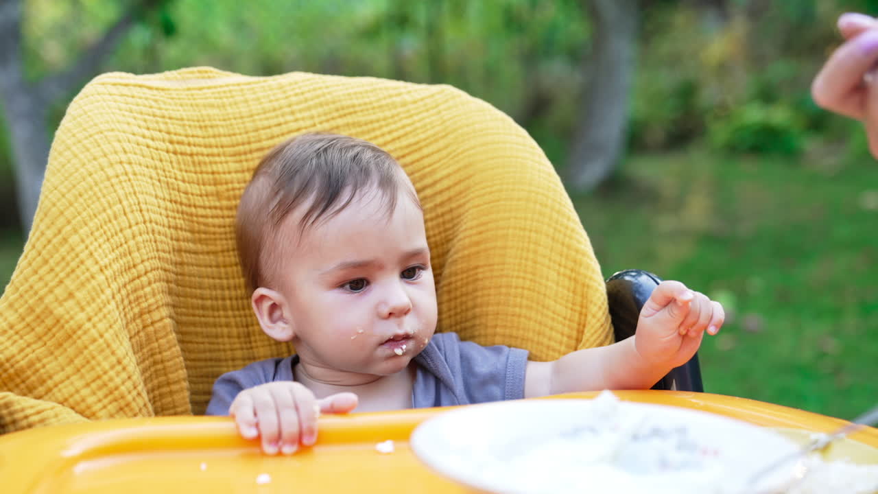 Adorable child at feeding table having lunch. Lovely kid looks at mom's hand filling spoon with porridge from a plate. Close up. Blurred backdrop.