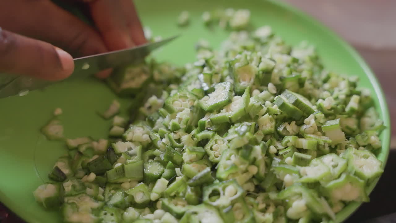 Tangan berkulit hitam mengiris okra di piring hijau, persiapan pasar cepat oleh penjual berkulit hitam menggunakan pisau untuk memotong okra siap jual, tampilan dekat sayuran bertekstur, tumpukan potongan, tempo energik.