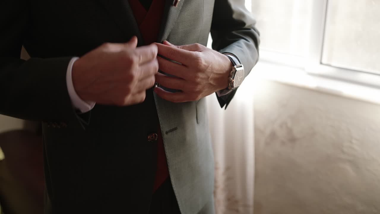 close up of groom buttoning jacket near window light