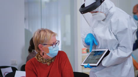 Woman-sitting-on-chair-listening-doctor-with-coverall-looking-on-tablet