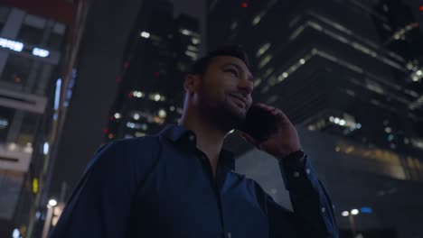 satisfied successful businessman calling with a smartphone outdoors, end the call and looking away with belief, standing by office buildings at night - orbit shot