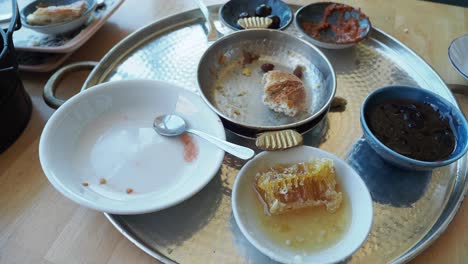 a close-up shot of an empty plate on a metal tray with a bowl of honey, a bowl of jam, some olives, and bread