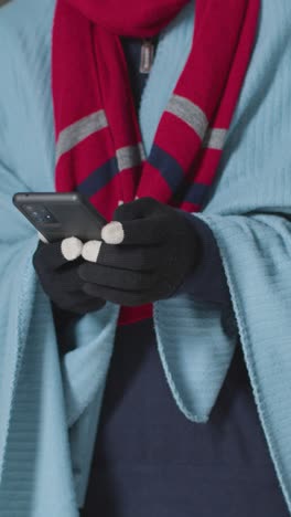 Vertical-Video-Close-Up-Of-Person-Wearing-Blanket-And-Gloves-Using-Mobile-Phone-Trying-To-Keep-Warm-In-Energy-Crisis