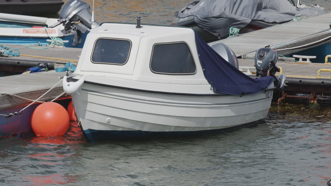 A white motorboat tied to a dock moves subtly with the water beside a bright orange buoy in a calm Scottish harbour under daylight