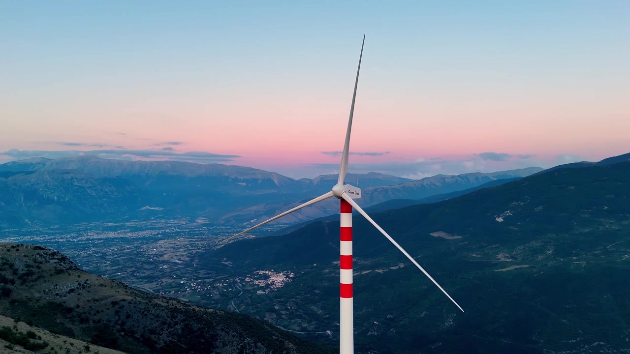 Drone view of a broken wind turbine standing motionless above Cocullo, Abruzzo, Italy, during blue hour, highlighting inactive renewable energy infrastructure against a dramatic Apennine mountain land
