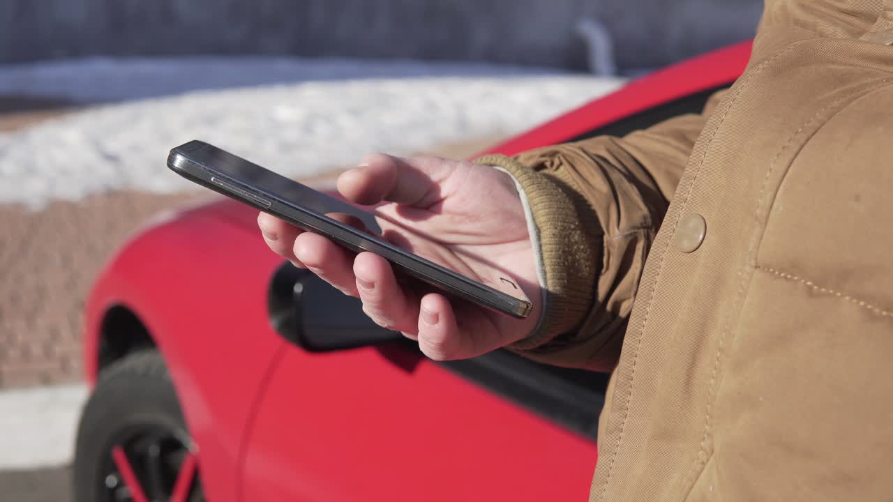 hombre joven sentado junto al coche, usando su teléfono inteligente, edificio moderno con grandes ventanas en el fondo