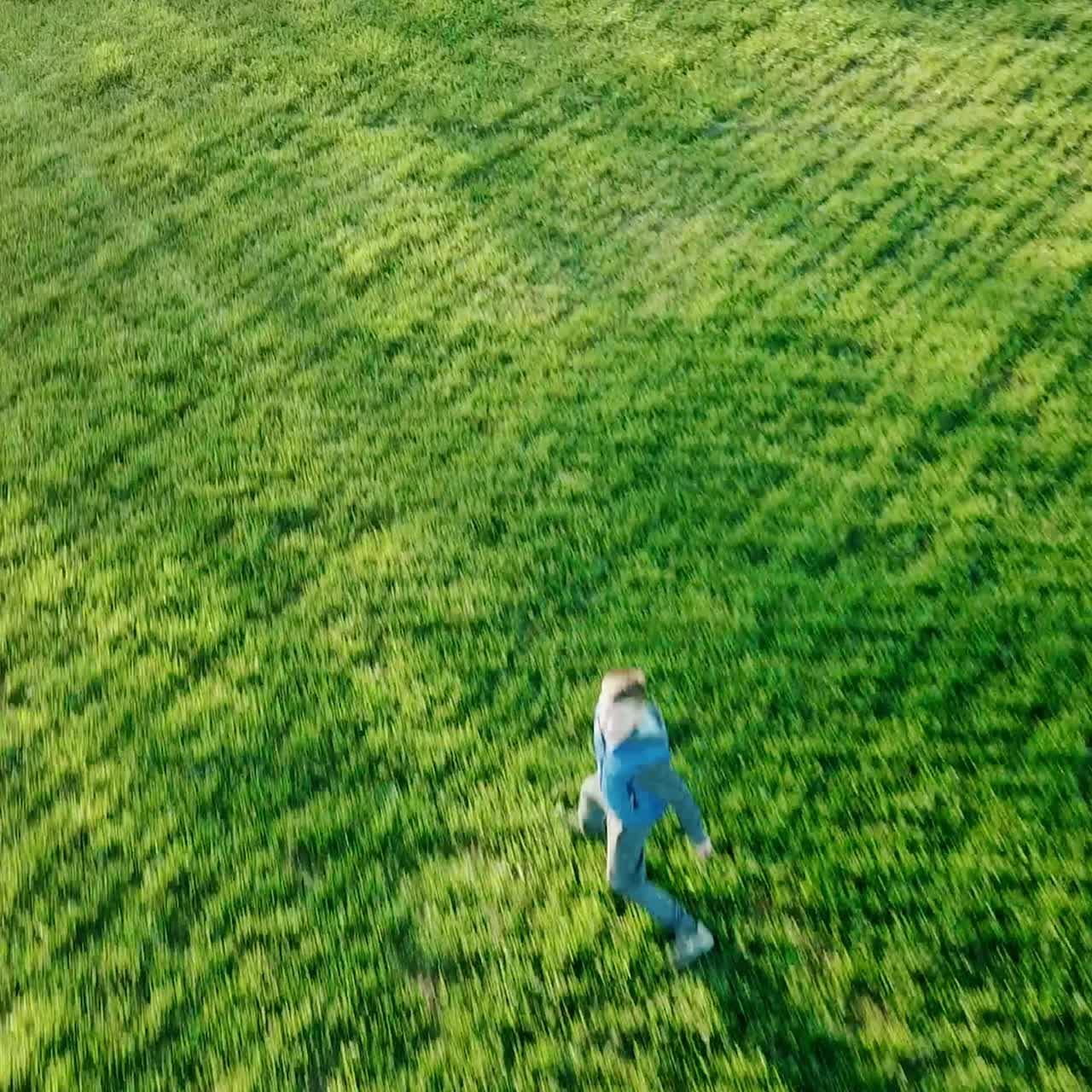 a little boy in a blue vest is running away to the river on the background of green field. Camera motion to right. Camera motion forward