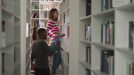 woman gives book to man in college library. librarian on step ladder serves reader with literature in bookstore. black student takes textbooks from worker