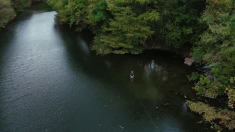 Texas-flyfisherman-casting-fly-rod-while-wading-in-a-beautiful-river-with-lush-tree-scenery-near-shore,-orbiting-spinning-aerial-drone-shot-in-4k