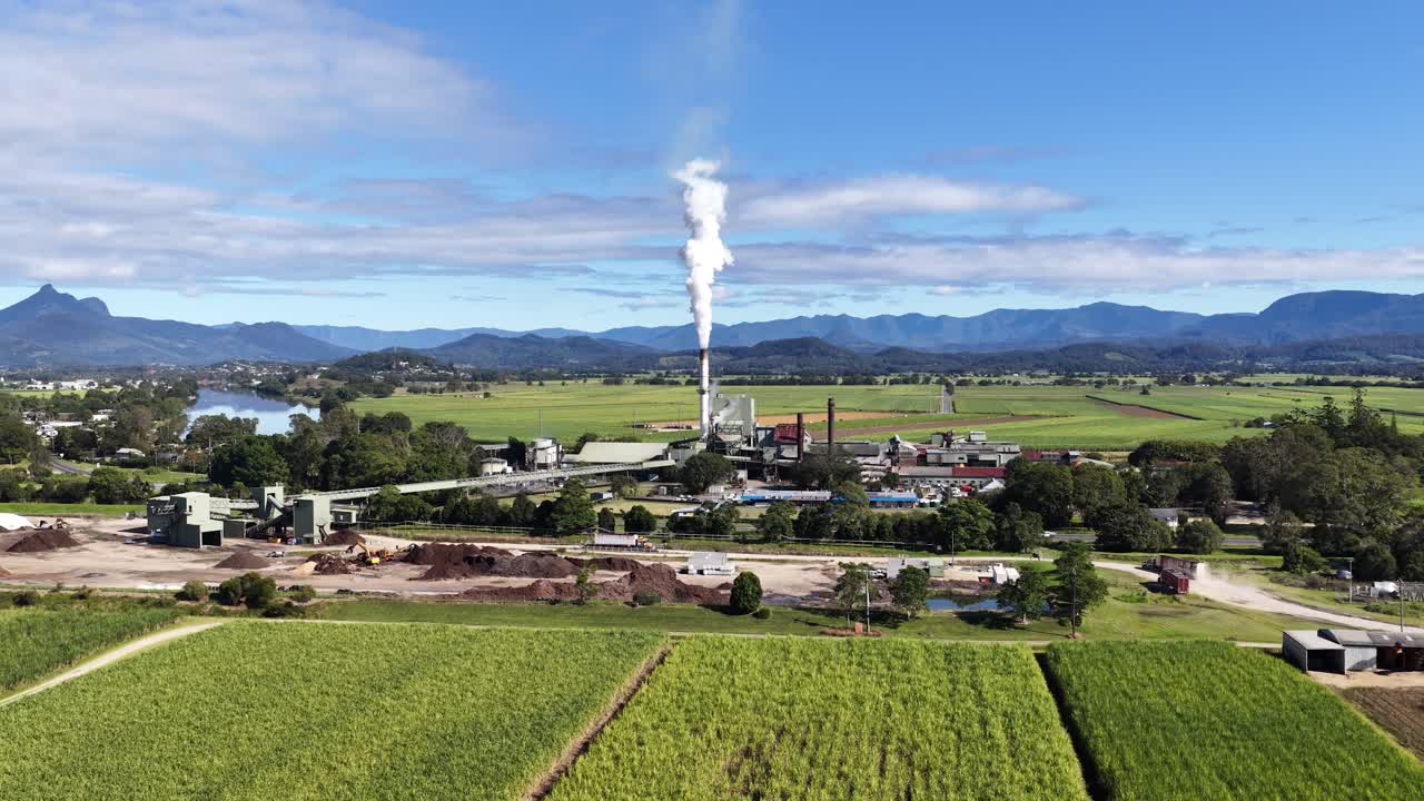 Aerial footage of a sugar mill with smoke rising, set against lush fields and distant mountains under clear skies