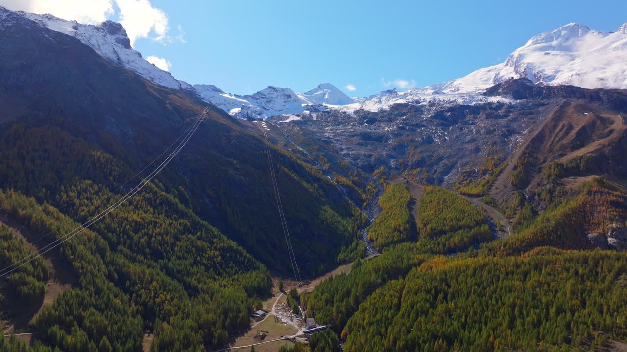 Stunning aerial footage of Saas-Fee in autumn. Golden forests surround the alpine village, nestled in the Swiss Alps. Vibrant fall colors and majestic peaks highlight the beauty of the season.