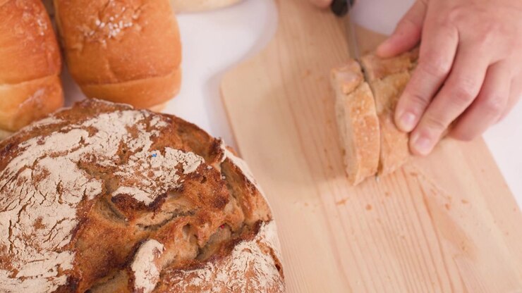 Close-up of hands slicing crusty bread on a wooden board amidst various bakery items.
