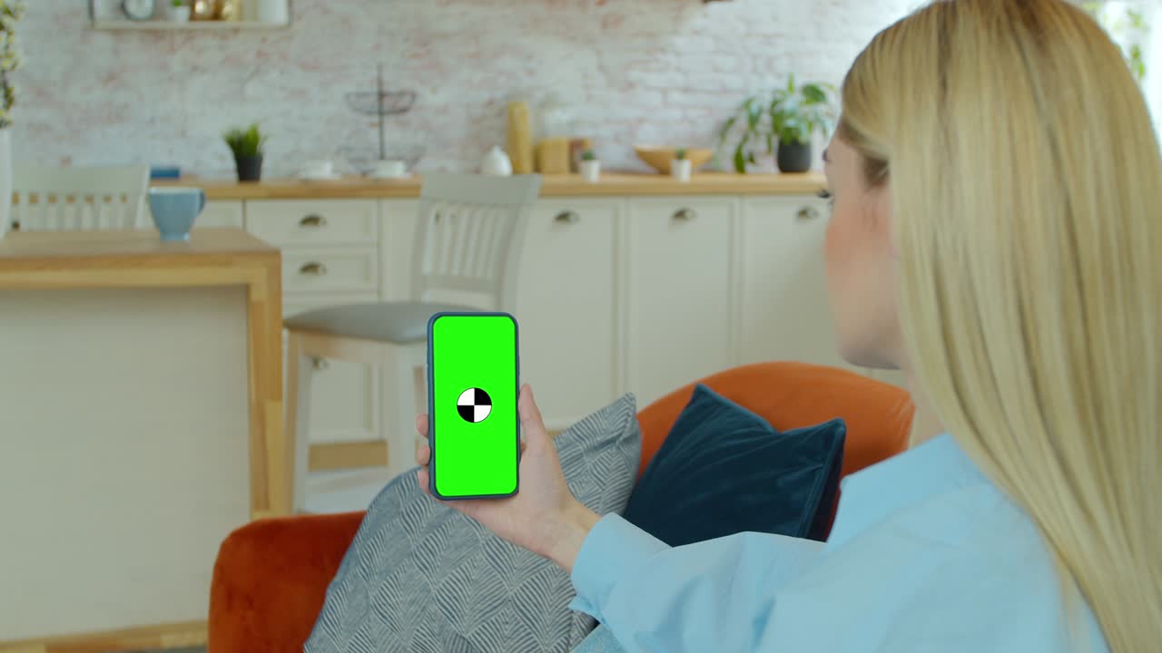 멋진 소녀가 스마트폰에서 비디오 통화를 하고 있습니다. 집에 있는 여자는 녹색 모형 화면 스마트폰을 사용합니다. 그녀는 그의 쾌적한 거실에서 소파에 앉아 있습니다.
