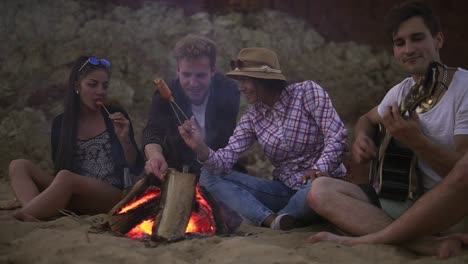 Group-Of-Young-And-Cheerful-People-Sitting-By-The-Fire-On-The-Beach-In-The-Evening,-Singing-Songs-And-Playing-Guitar