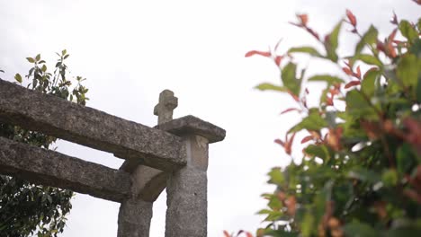 weathered granite cross atop rustic stone arch next to green leafy shrubs