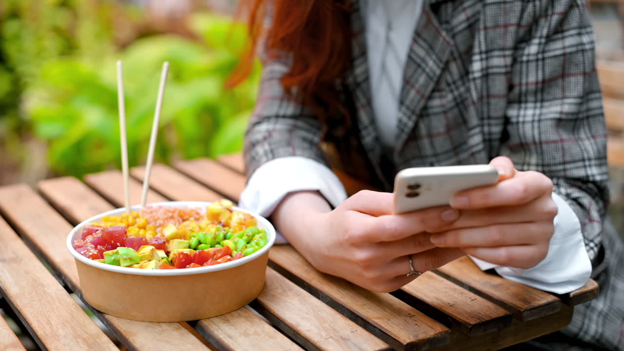 Close view of a woman using her phone, a poke bowl with vegetables, fruits and chopsticks on the table. Park with a lot of greenery. Slow motion