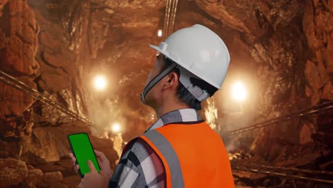 close up back view of asian male engineer with safety helmet working on a green screen smartphone and looking around while standing in underground mine tunnel