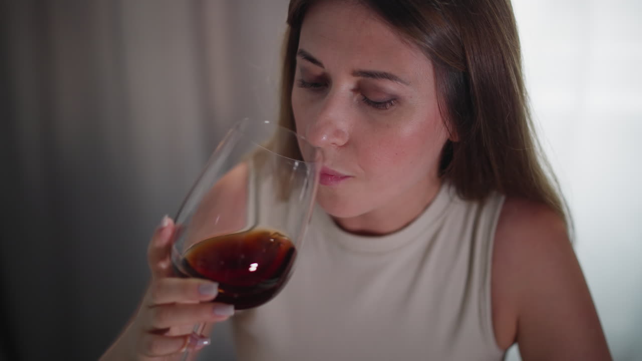 Close up of young woman in sleeveless top quickly drinking dark red wine from large glass, natural indoor lighting, focused expression, casual setting with soft background